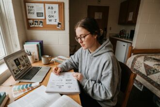 Étudiante en BTS design graphique travaillant dans un petit studio meublé économique, entourée de livres et de croquis sur son bureau compact